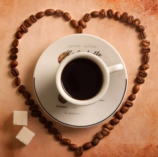 Herz aus Kaffeebohnen um Tasse auf altem Papier. Ansicht von oben — Stockfoto
