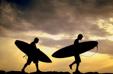 Two Surfers Carrying Their Boards Home At Sunset clipart