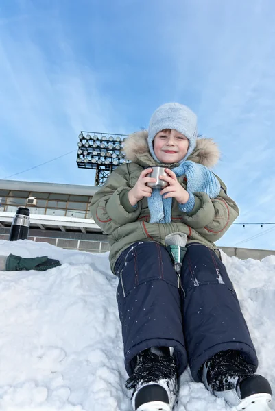 stock image The child with skates sits in a snowdrift and drinks tea