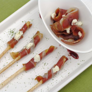 Thin bacon slices wrapped around thin salt sticks and served on a white rectangular plate on a green place mat