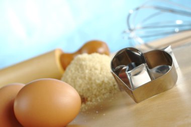 Heart shaped cookie cutter with baking ingredients and utensils such as eggs, brown sugar, rolling pin and a beater (Selective Focus, Focus on the front of the clipart