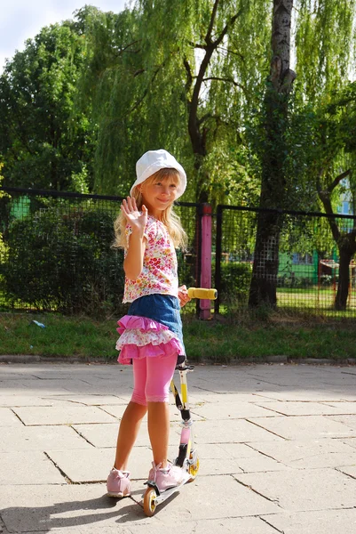 Young girl riding scooter and waving hand to friends — Stock Photo, Image