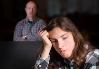 A troubled looking teenage boy with his father looking on in the background clipart