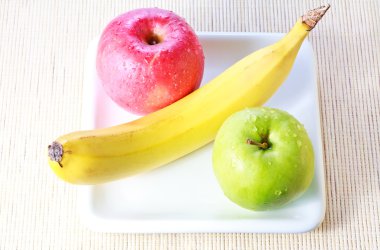 Banana, green and red apples on white plate over bamboo mat background clipart