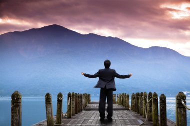 Businessman stand on the pier and watching the mountain and cloud of sunrise clipart