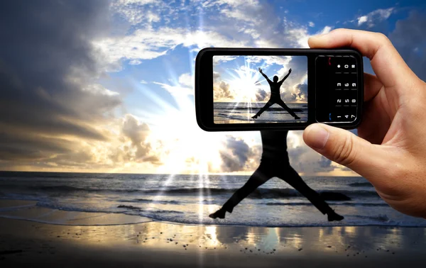 Cámara de teléfono móvil y feliz hombre saltando en la playa en el hermoso amanecer —  Fotos de Stock