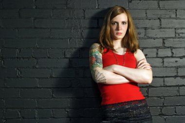 A young female with full arm tattoo leaning up against a black brick wall. clipart