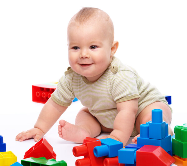 Cute little boy is playing with building bricks while sitting on floor, isolated over white