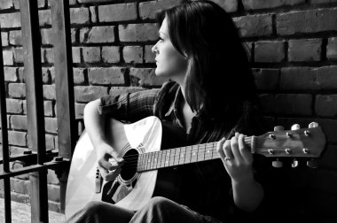 Tuğla duvara yaslanmış gitar çalan genç bir kadın parmaklıklardan içeri bakıyor..