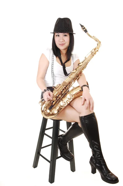 一名年轻亚裔女子坐在演播室里 吹奏萨克斯管穿短裤与吊杆和一顶帽子在她黑色的头发 为白色背景上 — 图库照片
