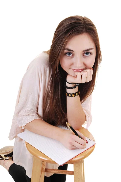 美丽的少年跪在一张椅子上 写在她的笔记本 微笑的相机 白色背景 — 图库照片