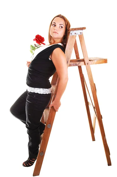 黒のズボンと白いシャツ彼女の手に赤いバラを保持 脚立の上に 明るい灰色の背景に対して傾いていると 素敵な若い女の子 — ストック写真