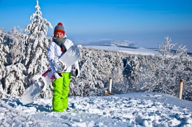 güzel bir kadın snowboard ile dağın zirvesinde duruyor. karla kaplı çam ağaçları ve mavi gökyüzü fonunda.