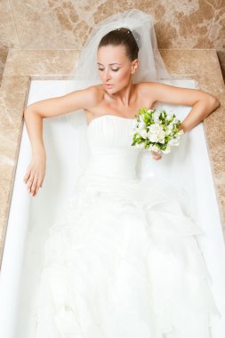 Attractive caucasian bride lying down in bath wearing wedding dress clipart