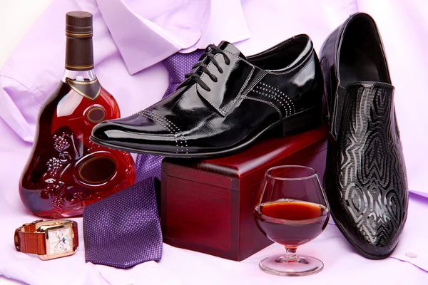 Комплект мужской обуви, бутылка и стакан бренди, а также мужские наручные часы, надетые на фиолетовую рубашку с галстуком — стоковое фото
