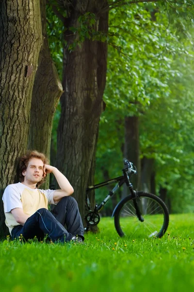 Ung man lokalisering i parken under trädet nära hans cykel — Stockfoto