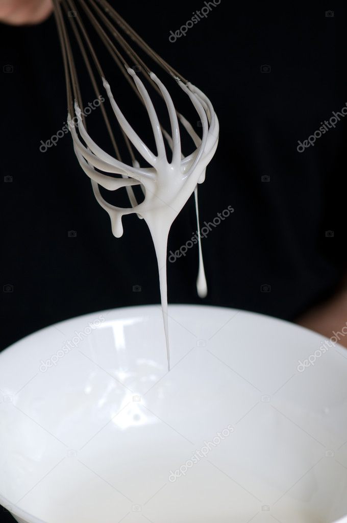 Whisk with creamy egg white close up over white bowl