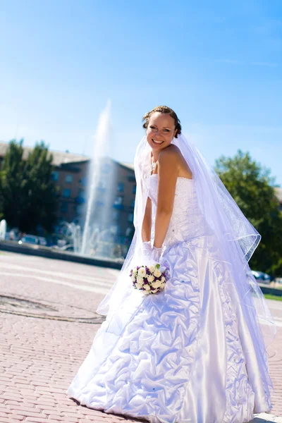 Beautiful bride in white dress holding bouquet and sat next to water founta — Stock Photo, Image