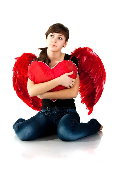 Beautiful girl sitting with heart shaped red pillow in red angel wings — Stock Photo, Image