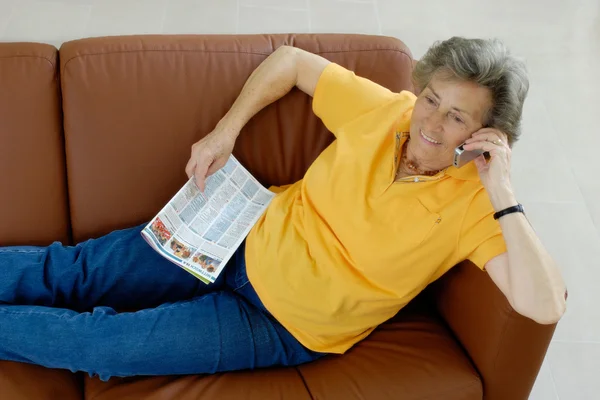 Ανώτερος γυναίκα με το tv-περιοδικό και το κινητό τηλέφωνό σας σε έναν καναπέ — Φωτογραφία Αρχείου
