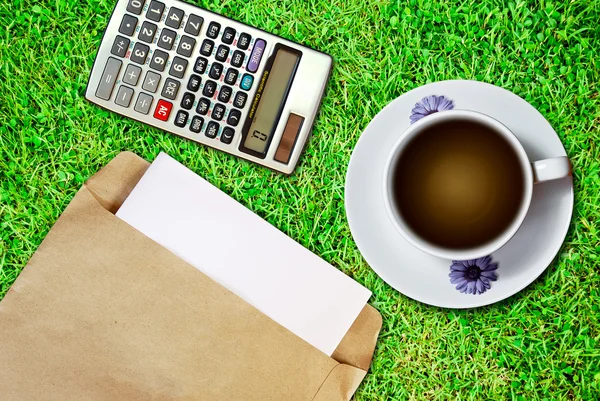 新鮮な緑の芝生の背景上の電卓でコーヒーと茶色の封筒文書の白いカップ — ストック写真