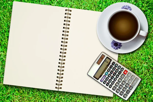 白いコーヒーのカップと新鮮な緑の芝生の背景に電卓を持つ空白のノートブック — ストック写真