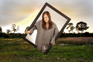 A lovely young brunette appears to be emerging from a second dimension through a black, wooden picture frame into a scenic outdoor location. clipart