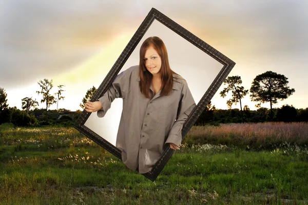 stock image A lovely young brunette appears to be emerging from a second dimension through a black, wooden picture frame into a scenic outdoor location.