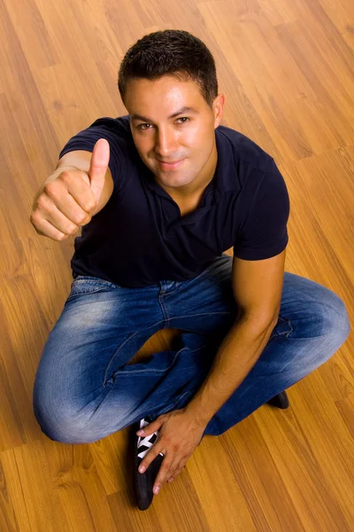 Молодой человек, сидящий на полу, поднимает большие пальцы — стоковое фото