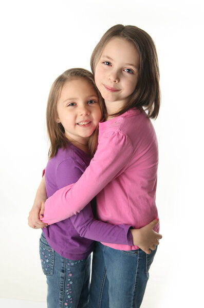 Two cute young sisters standing up hugging each other, isolated on white