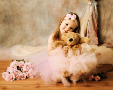 Adorable little girl dressed as a ballerina in a tutu, hugging a teddy bear sitting next to pink roses. clipart