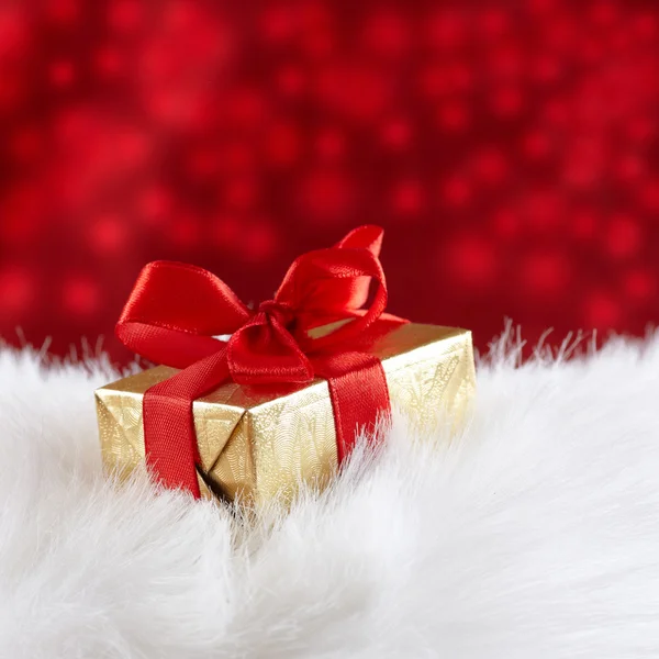 Golden gift box with red ribbon on white fur against red blurred — Stock Photo, Image