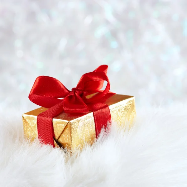 Golden gift box with red ribbon on white fur against silver blur — Stock Photo, Image
