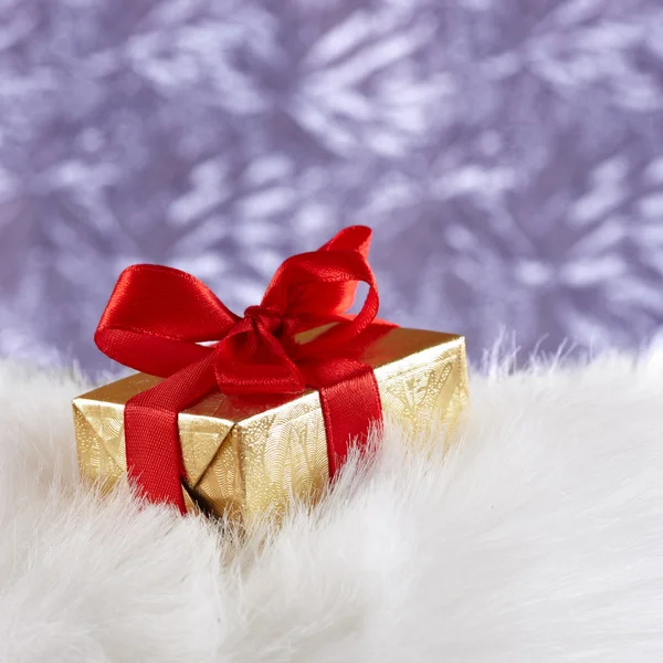 Golden gift box with red ribbon on white fur against blue blurre — Stock Photo, Image