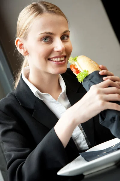 Bir sandviç yeme ve kameraya bakarak genç gülümseyen iş kadını — Stok fotoğraf