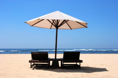 Two chaise lounges under an umbrella on sandy to an ocean coast clipart