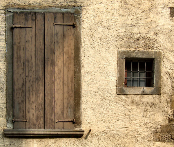Small window and big closed window with brown shutters. Rough, yellow or beige wall of an old house in a small Italian town or village.