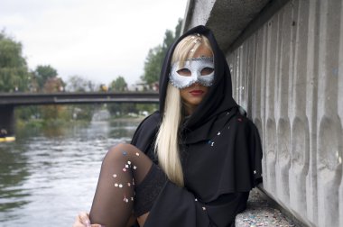 nehir kenarında oturan beyaz karnaval maskesi ve siyah pelerin ile seksi sarışın kız