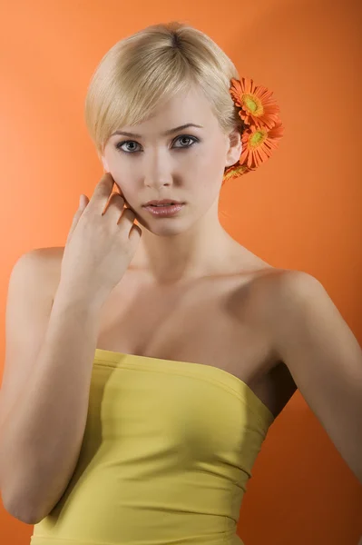 イエロー トップとガーベラ オレンジ色の背景上で作られた髪スタイルを持つ若い美しさブロンドの女の子 — ストック写真