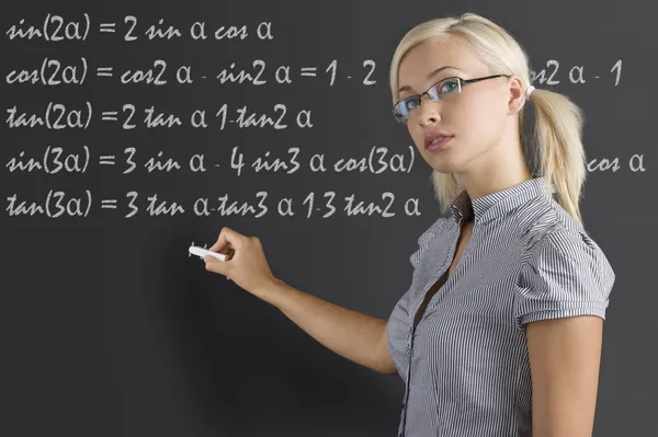 年轻漂亮老师与金色的头发和眼镜在黑板上考虑数学的一课 — 图库照片