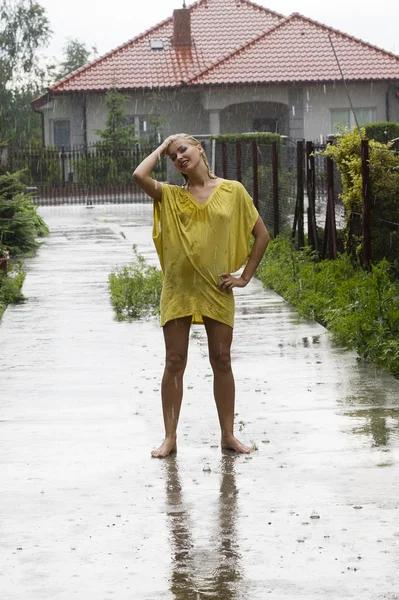 blond woman wearing a yellow dress just outside of her home under the rain falling down
