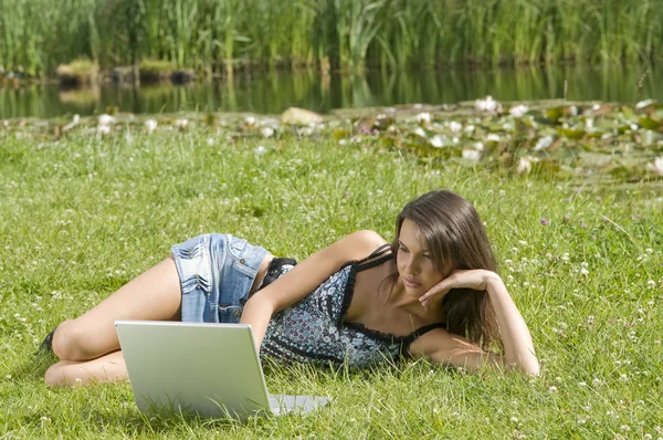 可爱布鲁内特穿便服躺在草地上和在计算机上工作 — 图库照片