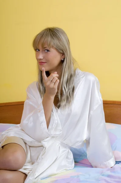 漂亮的金发女孩坐在床上使脸上的白色睡衣 — 图库照片