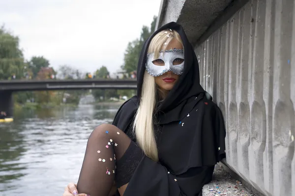 川の近く白いカーニバル マスクと黒マントと座っているセクシーなブロンドの女の子 — ストック写真