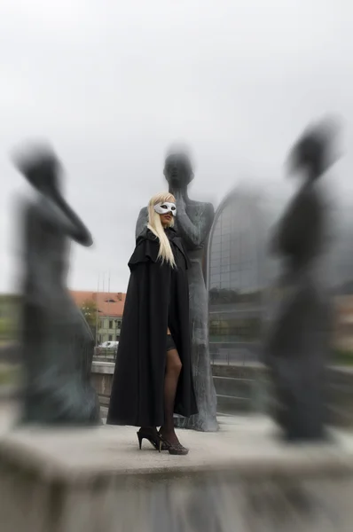 カーニバル マスクと黒マントを着ての大きな彫像の間ブロンドの女の子 — ストック写真