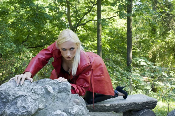 赤い皮のコートと岩の上に座って公園の外ブロンドのセクシーな女性 — Φωτογραφία Αρχείου