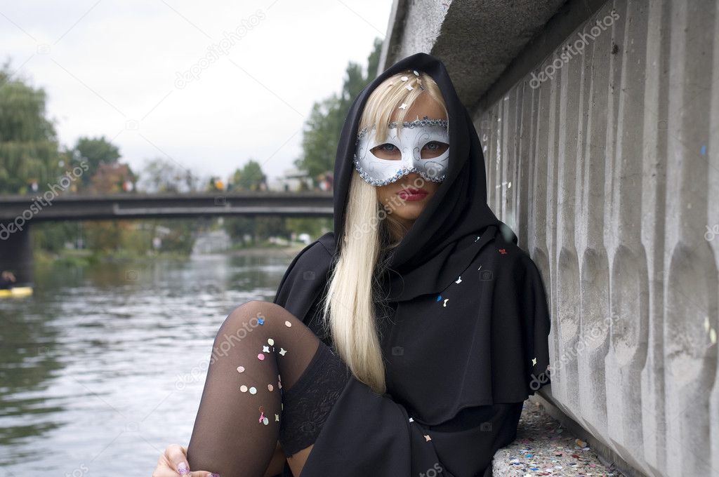 sexy blond girl sitting near river with white carnival mask and black cloak