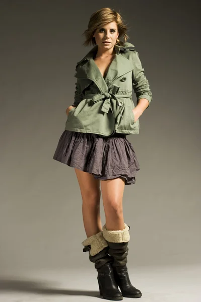 灰色の背景上の緑のコートとミニのスカートを着て美しいブロンドの女の子 — ストック写真