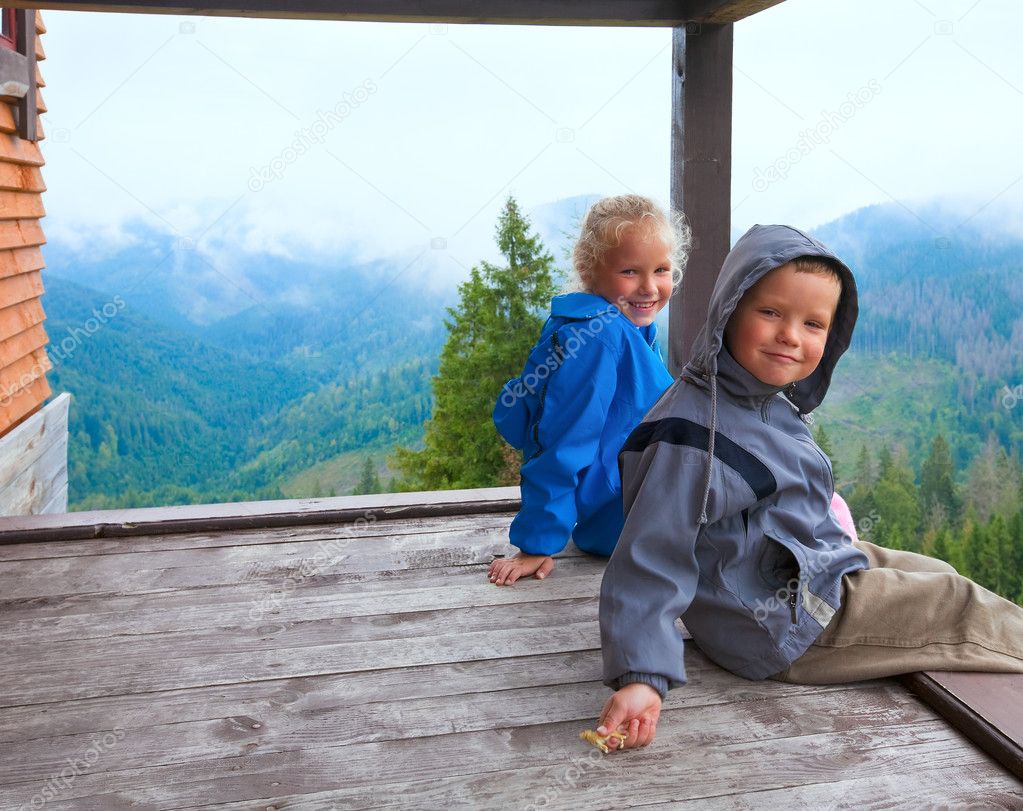 Small boy and girl on wooden cottage porch on mountain hill top and cloudy morning view behind