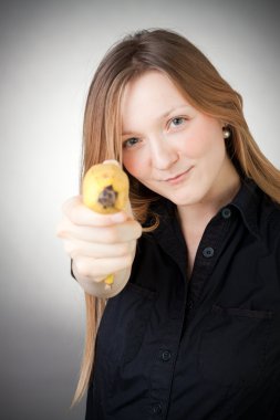 Pretty girl in a black dress is holding a banana as a gun, with grey background clipart